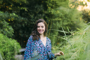 Smiling beautiful woman standing amidst plants in park - DCRF00855