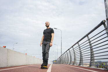 Lächelnder junger Mann, der beim Inlineskaten auf einer Brücke vor bewölktem Himmel wegschaut - VPIF03039