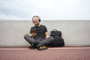 Junger Mann auf Inline-Skates, der sein Smartphone benutzt, während er mit seinem Rucksack auf einer Brücke sitzt - VPIF03049