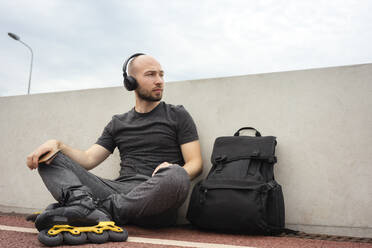 Thoughtful young man wearing inline skates listening music while sitting on bridge - VPIF03050
