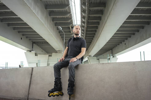 Nachdenklicher junger Mann auf Inline-Skates, der auf einer Stützmauer unter einer Brücke sitzt - VPIF03051