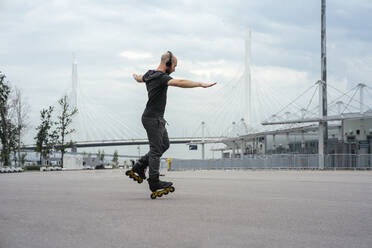 Man with arms outstretched listening music while inline skating on road against cloudy sky - VPIF03059