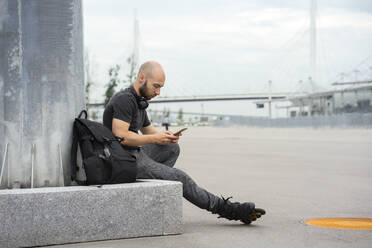 Junger Mann in Rollschuhen, der ein Mobiltelefon benutzt, während er auf einem Sitz in der Stadt sitzt - VPIF03062