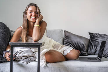 Lächelnde Frau mit Smartphone sitzt zu Hause auf der Couch - DLTSF01142
