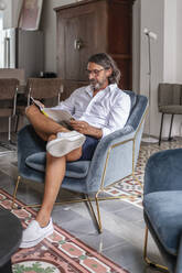 Man reading book while sitting on armchair at home - DLTSF01143