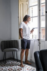 Mann mit Händen in den Taschen, der sein Smartphone hält, während er durch das Fenster seines Hauses schaut - DLTSF01144