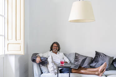 Lächelnder Mann, der einen Laptop benutzt, während er zu Hause auf der Couch sitzt - DLTSF01160