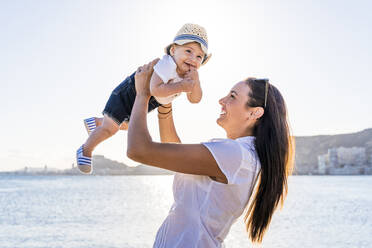 Happy mother picking up son while standing against sea at sunset - DLTSF01189