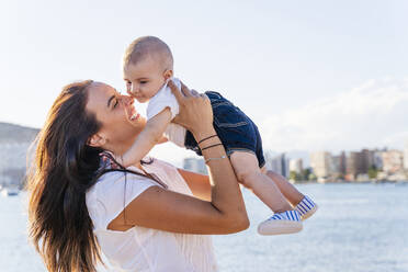 Smiling mother picking up baby son while standing against sea at sunset - DLTSF01190