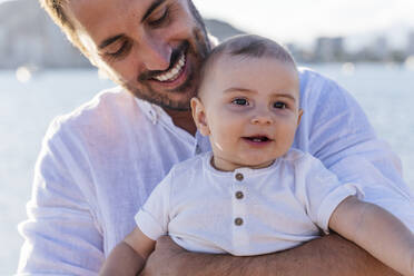 Close-up of smiling father carrying cute son at beach during sunset - DLTSF01192