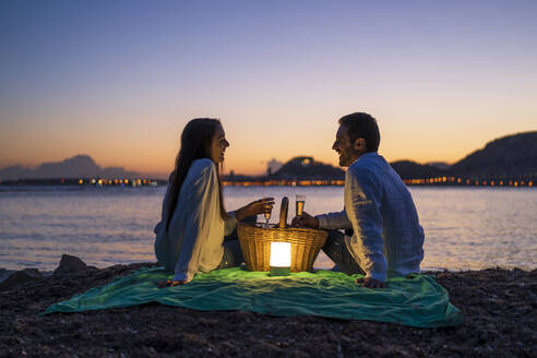 Paar mit Getränken, die sich gegenseitig ansehen, während sie in der Abenddämmerung am Strand gegen den klaren Himmel sitzen - DLTSF01211