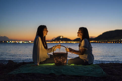Schwestern mit Getränken im Gespräch, während sie auf einer Decke am Strand gegen den klaren Himmel in der Abenddämmerung sitzen - DLTSF01214