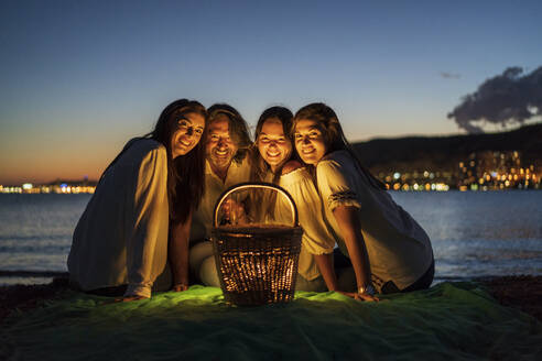 Älterer Mann mit Töchtern auf einer Decke sitzend gegen das Meer in der Abenddämmerung - DLTSF01216