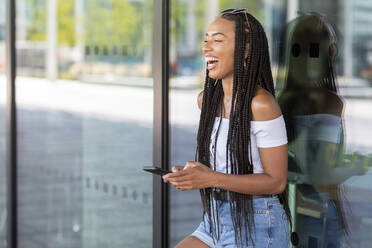 Frau lacht, während sie ein Mobiltelefon gegen eine Glaswand in der Stadt benutzt - WPEF03348
