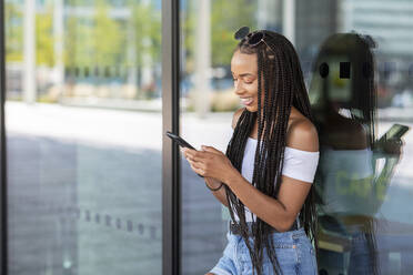 Woman using phone while leaning on glass wall in city - WPEF03349