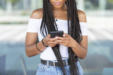 Junge Frau benutzt ein Mobiltelefon, während sie gegen eine Glaswand in der Stadt steht - WPEF03351