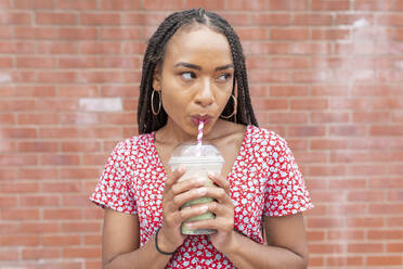 Young woman drinking smoothie while standing against brick wall - WPEF03386