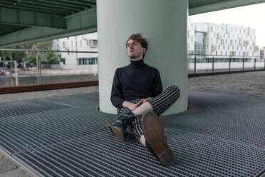 Thoughtful young man looking away while sitting by column on footpath - OGF00557