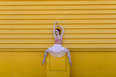 Smiling ballerina with arms raised dancing on seat against yellow wall - TCEF01065