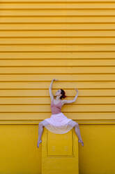 Junge Frau mit erhobenen Armen tanzt auf einem Sitz vor einer gelben Wand - TCEF01066