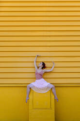 Young woman with arms raised dancing on seat against yellow wall - TCEF01066