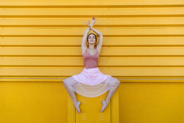 Selbstbewusste junge Frau mit erhobenen Armen tanzt Ballett auf einem Sitz vor einer gelben Wand - TCEF01067