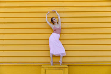 Ballerina with arms raised posing while standing on seat against yellow wall - TCEF01071