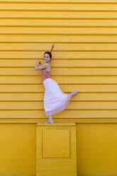 Smiling ballet dancer dancing on seat by yellow wall - TCEF01074
