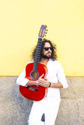 Man holding guitar while standing against yellow wall - JCMF01400