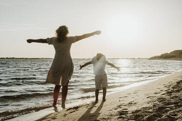 Grandmother and grandson with arms outstretched standing at beach during sunset - ERRF04343