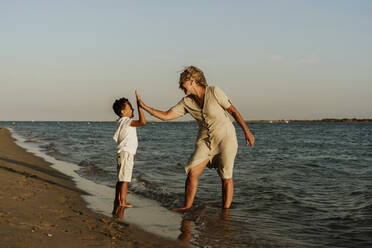 Happy grandmother and grandson doing high five to each other at beach during sunset - ERRF04344
