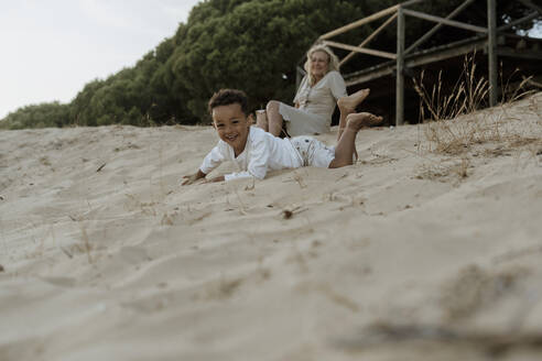 Großmutter sitzt, während der Enkel am Strand im Sand liegt - ERRF04360