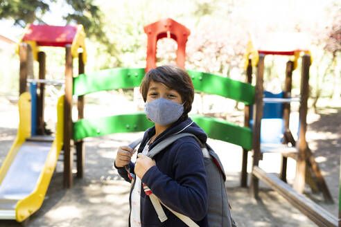 Boy wearing mask with backpack standing in schoolyard - VABF03463