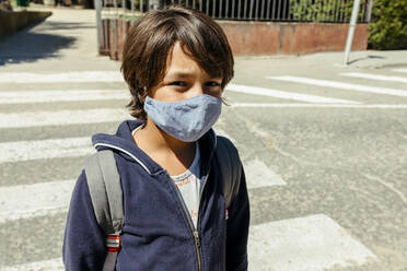 Schoolboy wearing mask standing on road during sunny day - VABF03466