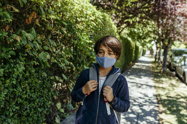 Schuljunge mit Maske, der wegschaut, während er bei Pflanzen auf dem Gehweg steht - VABF03469