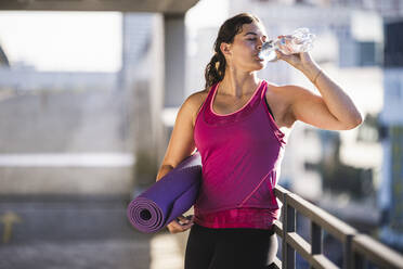 Junge Frau, die eine Gymnastikmatte hält und Wasser trinkt, während sie an einem Geländer auf einer Terrasse steht - UUF21363