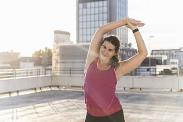 Young woman with arms raised exercising on terrace against sky at sunset - UUF21387