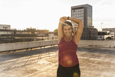 Smiling woman with arms raised exercising on terrace against sky at sunset - UUF21388