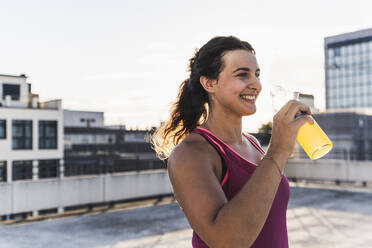 Smiling young woman having drink while standing on terrace against sky at sunset - UUF21391