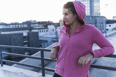 Nachdenkliche Sportlerin mit Kapuze, die bei Sonnenuntergang auf einer Terrasse am Geländer steht - UUF21394