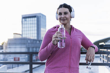 Junge Frau hält Wasserflasche und hört Musik über Kopfhörer auf einer Terrasse bei Sonnenuntergang - UUF21396