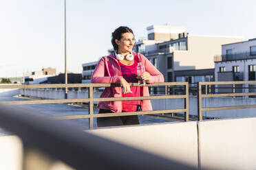 Nachdenkliche junge Frau mit Wasserflasche am Geländer einer Terrasse bei Sonnenuntergang - UUF21399