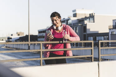 Junge Frau mit Wasserflasche und Smartphone auf einer Terrasse bei Sonnenuntergang - UUF21400