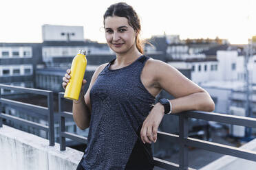 Lächelnde Sportlerin, die eine Flasche in der Hand hält, während sie an einem Geländer auf einer Terrasse steht - UUF21406