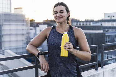 Young woman with eyes closed holding bottle while standing by railing on terrace - UUF21407