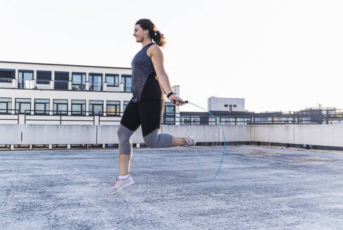 Junge Frauen trainieren mit Springseil auf der Terrasse gegen den klaren Himmel bei Sonnenuntergang - UUF21409