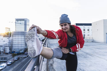 Young woman stretching leg on railing while standing on terrace against clear sky - UUF21414