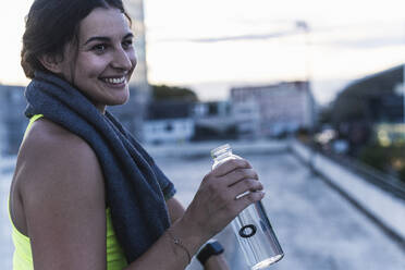 Nahaufnahme einer lächelnden jungen Frau, die eine Wasserflasche hält und wegschaut, während sie auf einer Terrasse sitzt - UUF21421