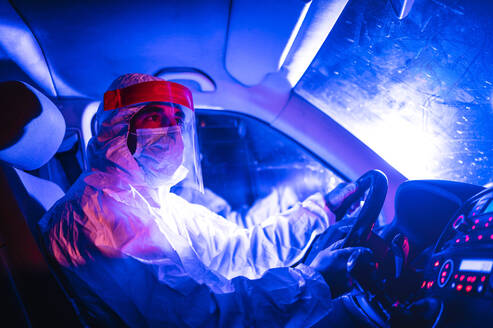 Male worker wearing protective suit driving ambulance at night - JCMF01418