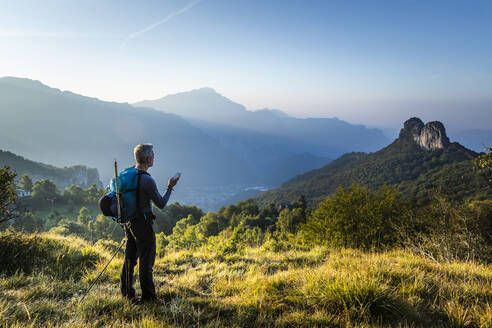 Mann mit Rucksack und Mobiltelefon bei Sonnenaufgang auf einem Berg stehend, Orobie, Lecco, Italien - MCVF00588
